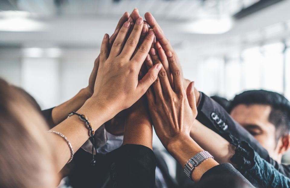 A group of coworkers in an office placing their hands together in the air to signify a huddle